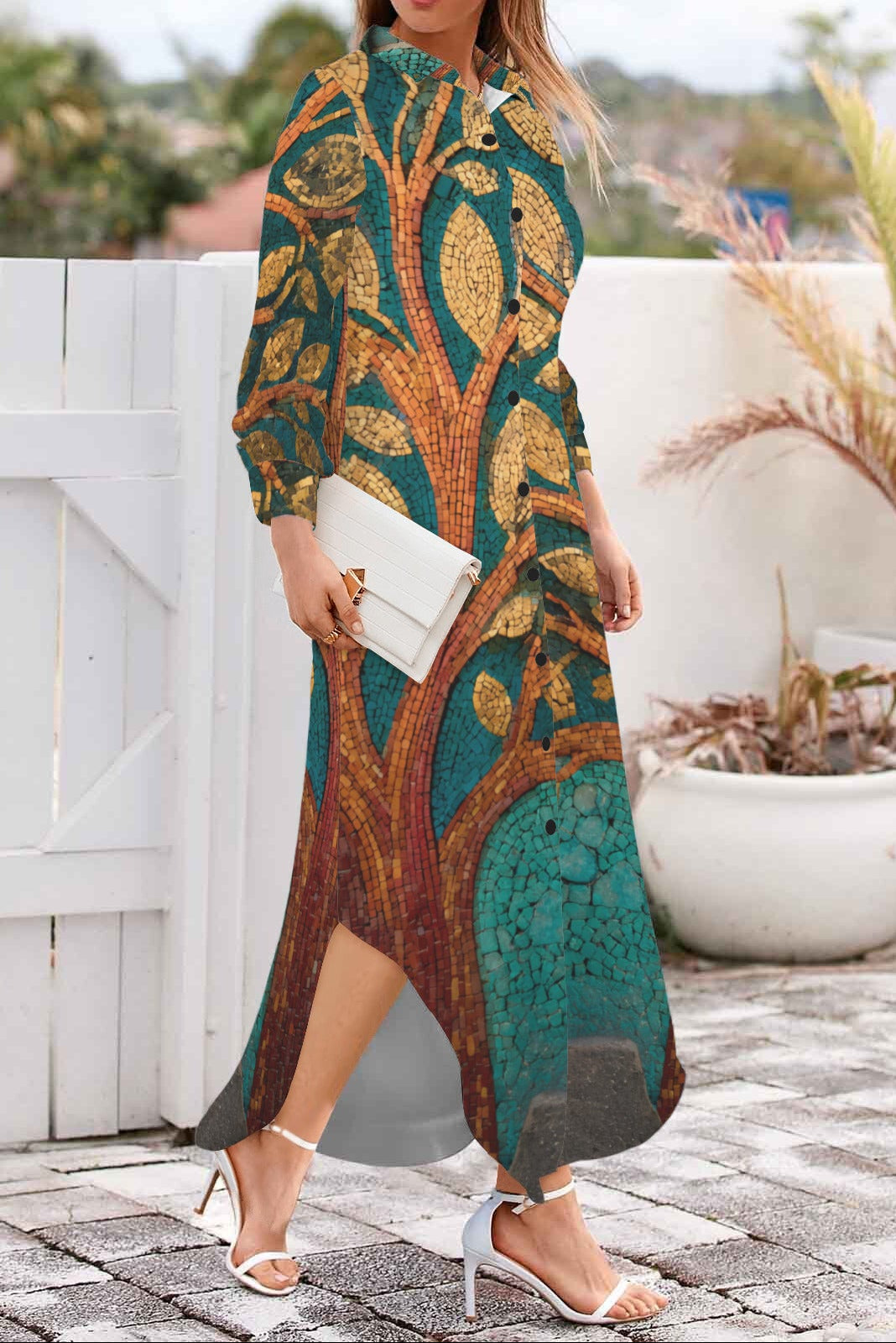 A woman wearing a long-sleeved maxi dress with a detailed tree trunk and leaf pattern in teal, gold, and brown, holding a white clutch and wearing white high-heeled sandals, standing on a stone patio in front of a white fence.