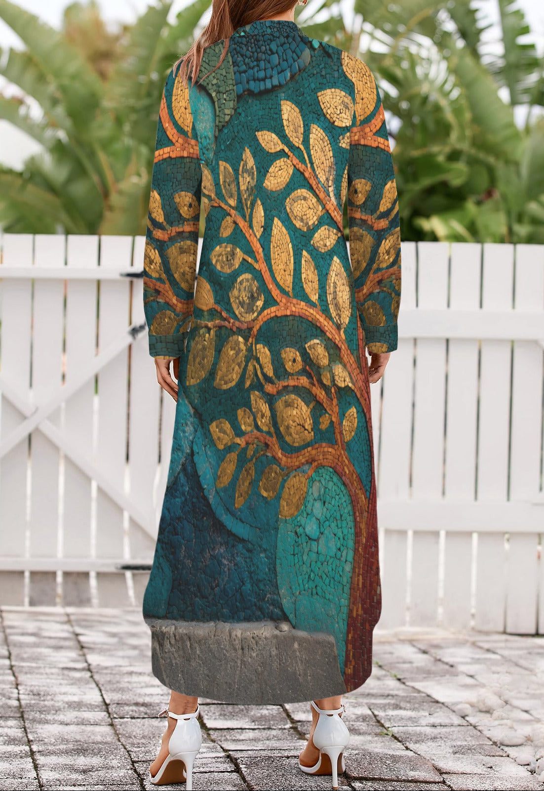 A woman viewed from behind wearing a long, loose-fitting maxi dress with a detailed tree branch pattern in teal, gold, and brown. She is standing on a stone patio in front of a white wooden fence with green foliage in the background.