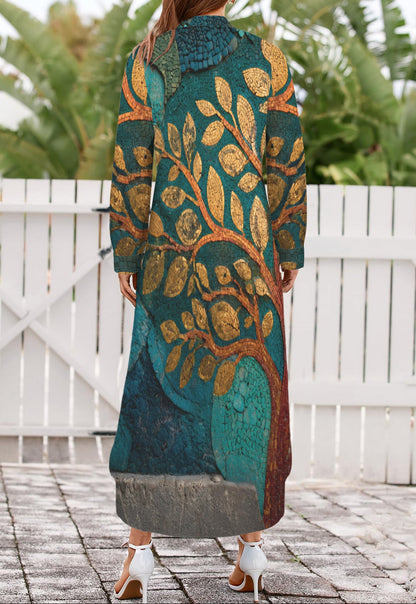 A woman viewed from behind wearing a long, loose-fitting maxi dress with a detailed tree branch pattern in teal, gold, and brown. She is standing on a stone patio in front of a white wooden fence with green foliage in the background.