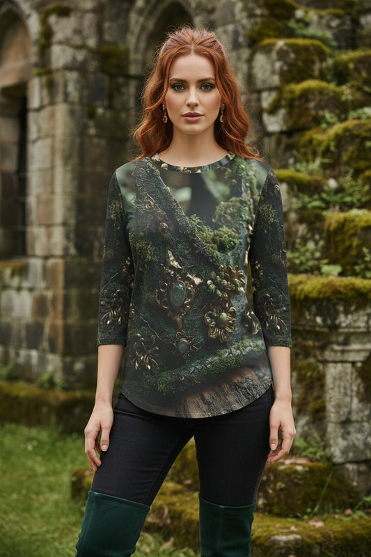 A woman with red hair stands outdoors in front of mossy stone ruins, wearing a three-quarter sleeve top with a dark forest and tree print, black leggings, and dark green boots.