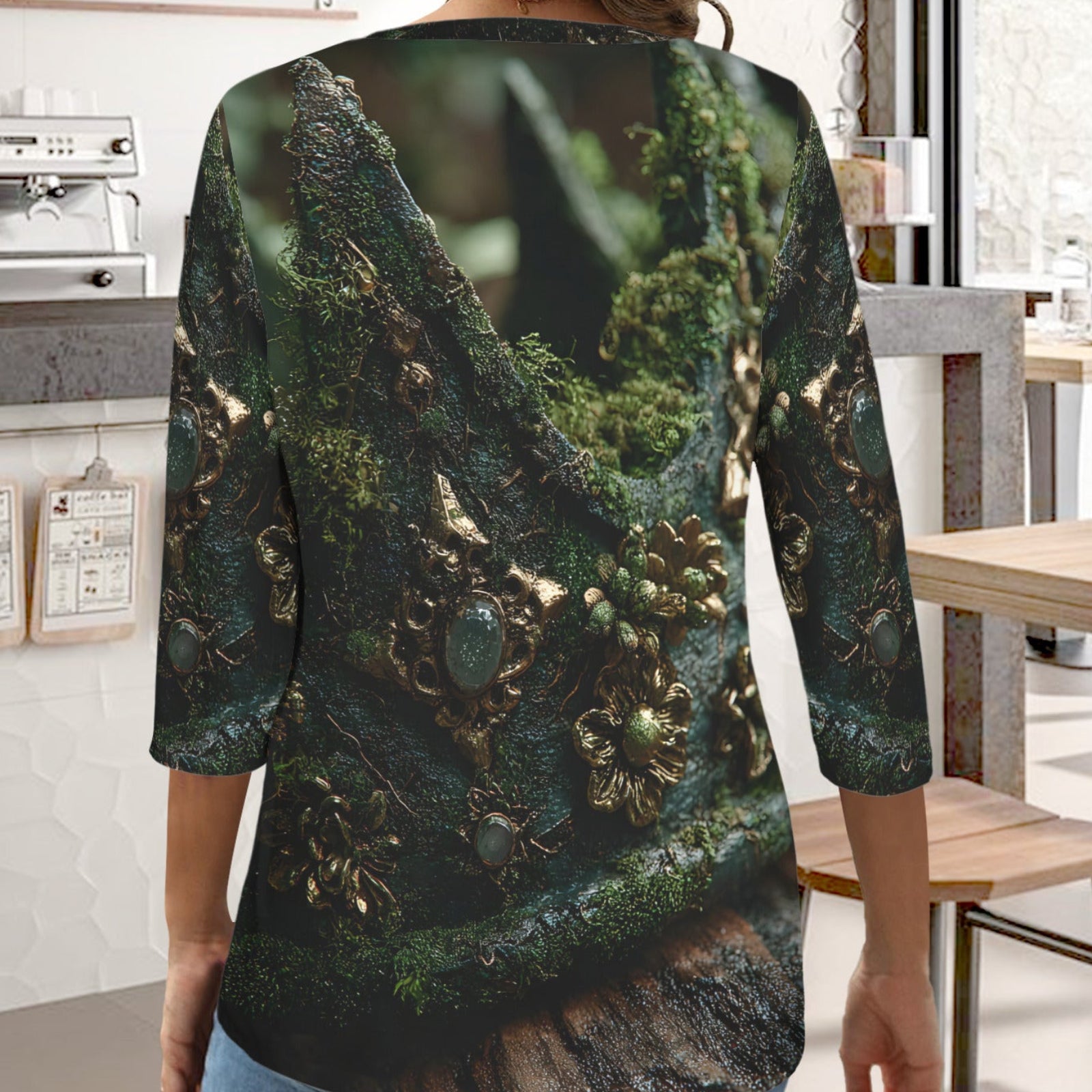 A woman wearing a three-quarter sleeve long-sleeve top with an all-over print of mossy tree roots and gold leaf details, shown from the back in a cafe setting.
