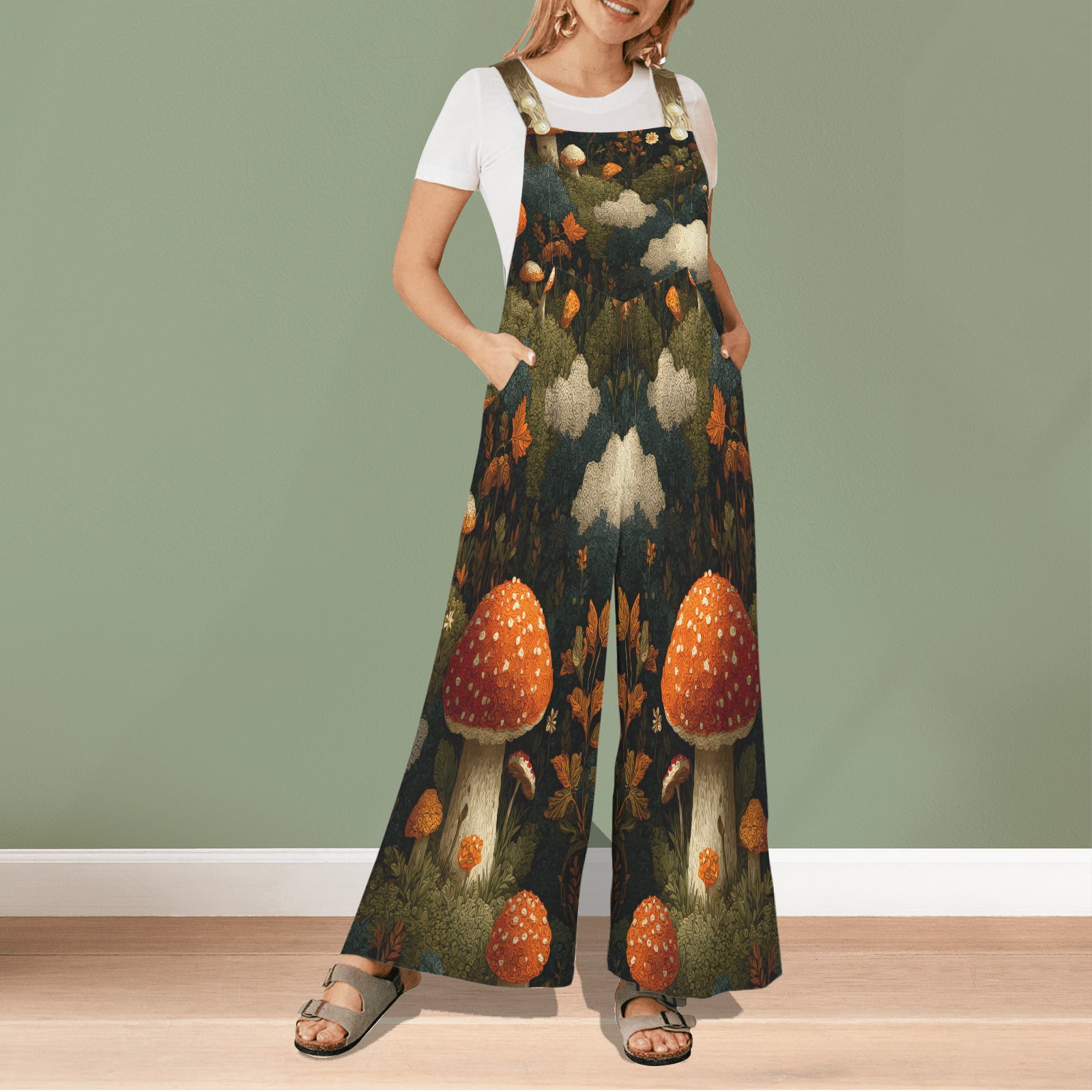 A woman modeling a pair of wide-leg jumpsuit overalls with a detailed forest and mushroom print, worn over a white t-shirt. The jumpsuit features a dark forest background with large orange and white mushrooms, green foliage, and beige clouds.