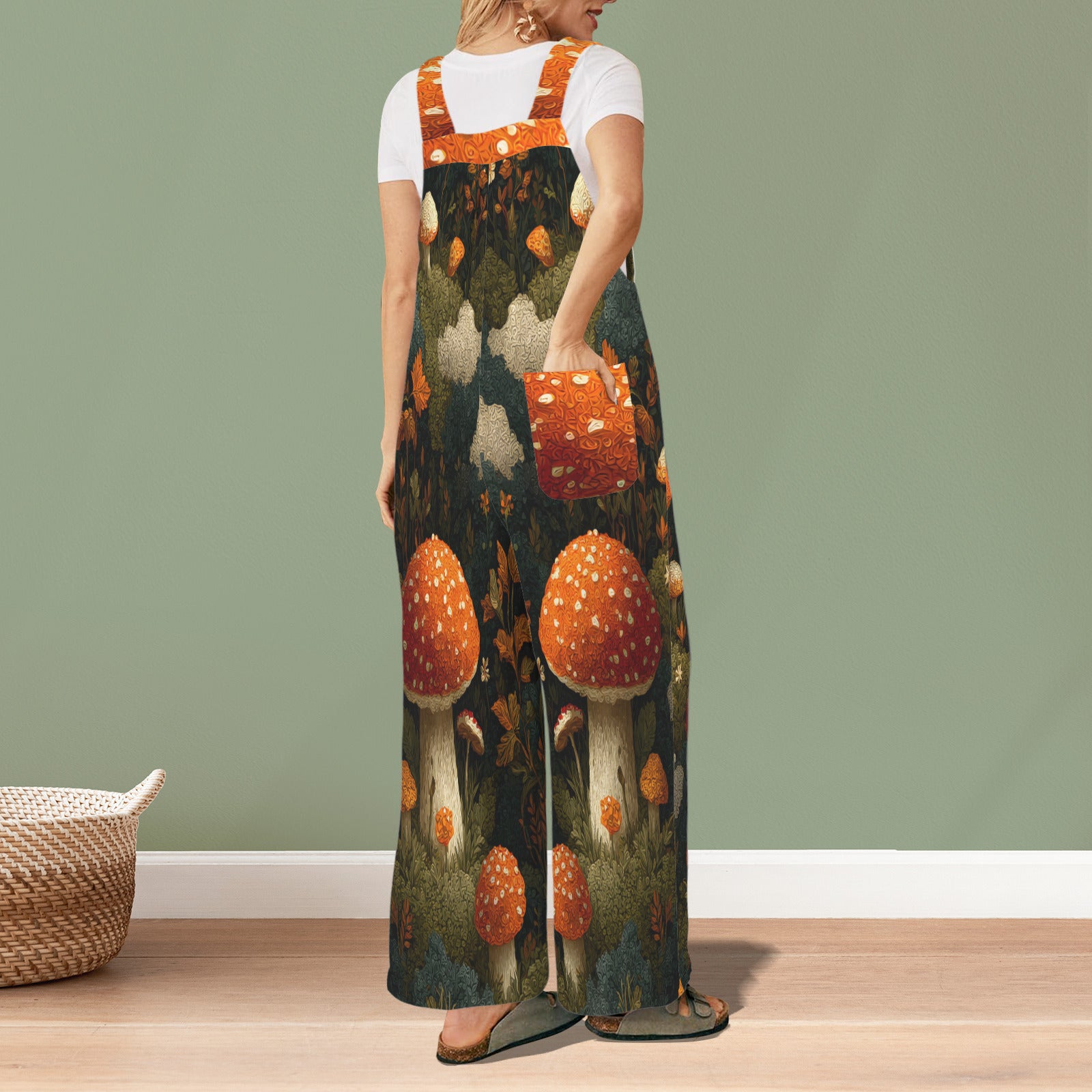 A woman stands in a room wearing a long-sleeve maxi overall dress with a whimsical forest mushroom print. The dress features orange and white spotted mushrooms on a dark forest background, worn over a white t-shirt.