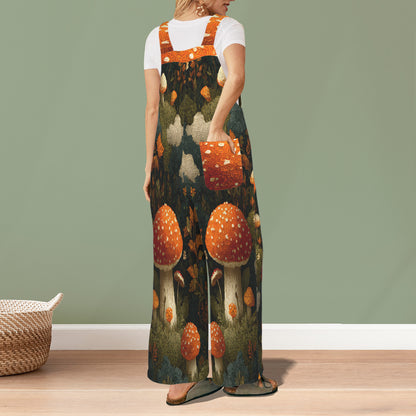 A woman stands in a room wearing a long-sleeve maxi overall dress with a whimsical forest mushroom print. The dress features orange and white spotted mushrooms on a dark forest background, worn over a white t-shirt.