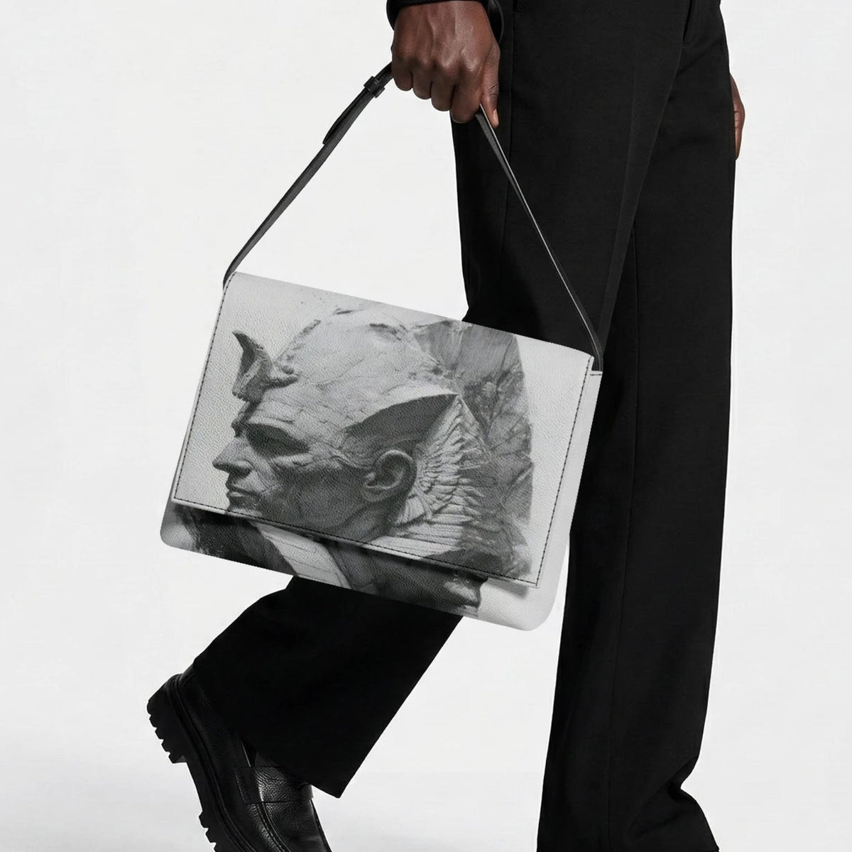 A person in black clothing carries a rectangular shoulder bag with a black-and-white sculptural face print on a light gray background, photographed against a clean white studio backdrop.