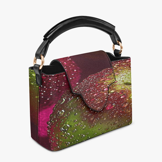 A structured handbag with a black top handle and gold-tone hardware, featuring a vibrant, abstract watercolor print in shades of magenta, green, and brown with a water droplet pattern, shown on a light gray background.
