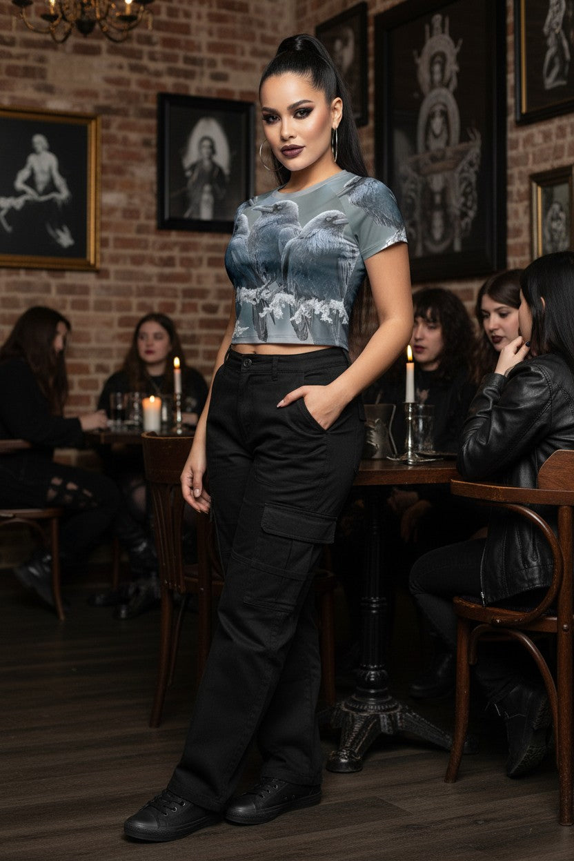 A woman stands in a dimly lit gothic-themed restaurant or cafe, wearing a short-sleeve cropped graphic top with a blue and gray gothic print, paired with black cargo pants and black shoes.