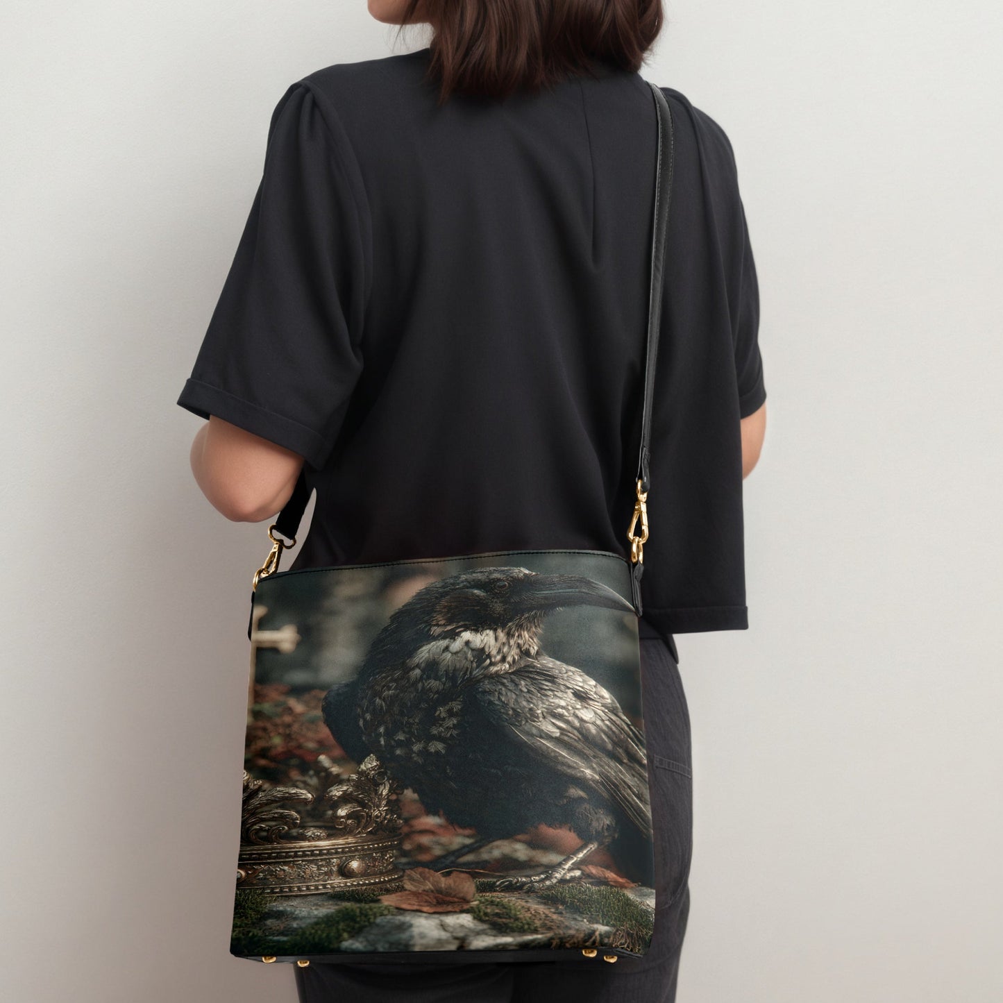 A person wearing a black t-shirt and dark pants, shown from the back while carrying a large shoulder bag. The bag features a detailed graphic print of a raven perched on a stone surface with a crown, and is accented with gold-tone hardware and a black strap.