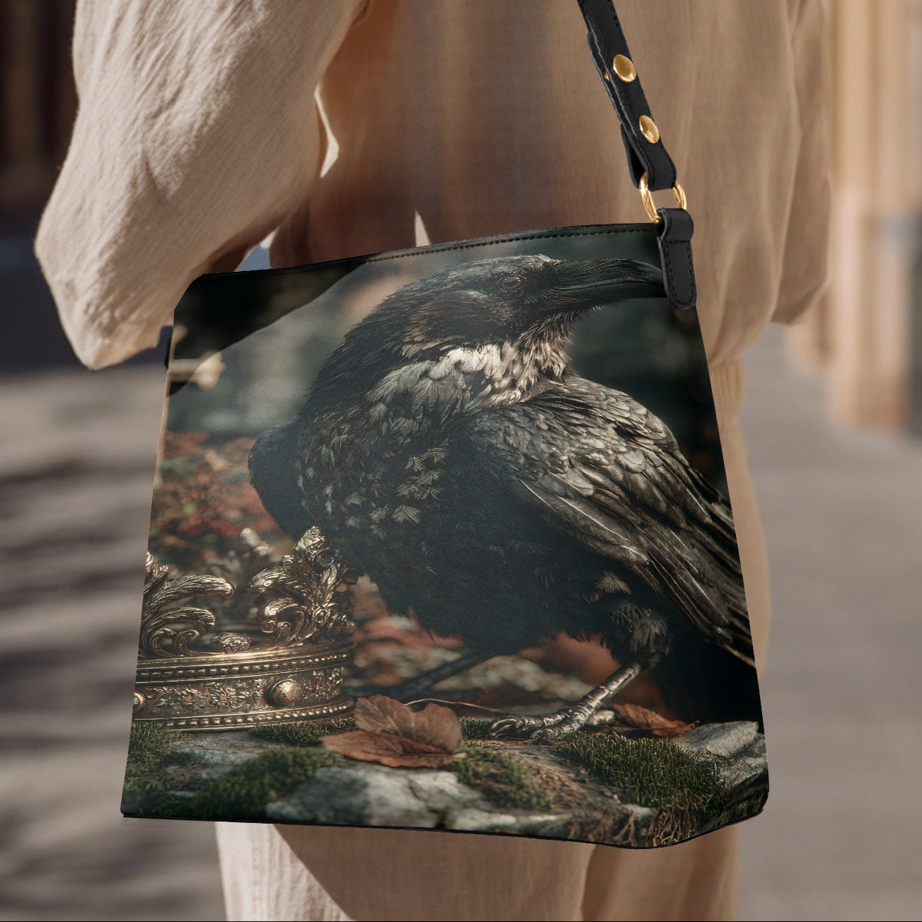 A woman wearing a cream-colored linen top carries a black shoulder bag featuring a detailed graphic print of a raven perched on a stone wall with a gold crown.