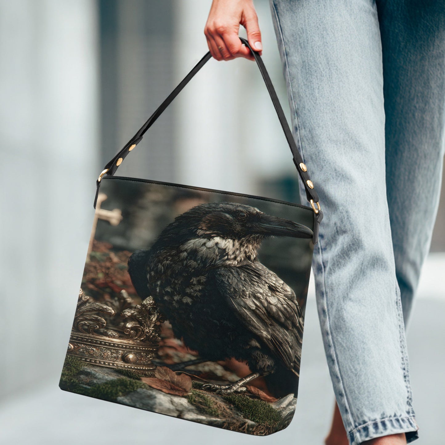 A person in light-wash jeans carries a shoulder bag featuring a printed design of a dark raven perched on a stone surface with autumn leaves.
