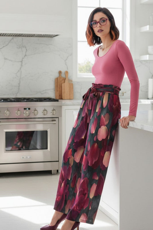 A woman wearing a pink long-sleeve top and a dark floral wide-leg skirt with purple and pink tones, standing in a bright modern kitchen.