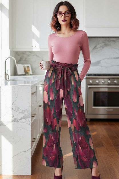 A woman stands in a bright, modern kitchen wearing a dusty pink long-sleeve top and high-waisted, wide-leg trousers with a dark red and green floral pattern. She is holding a mug and wearing glasses.