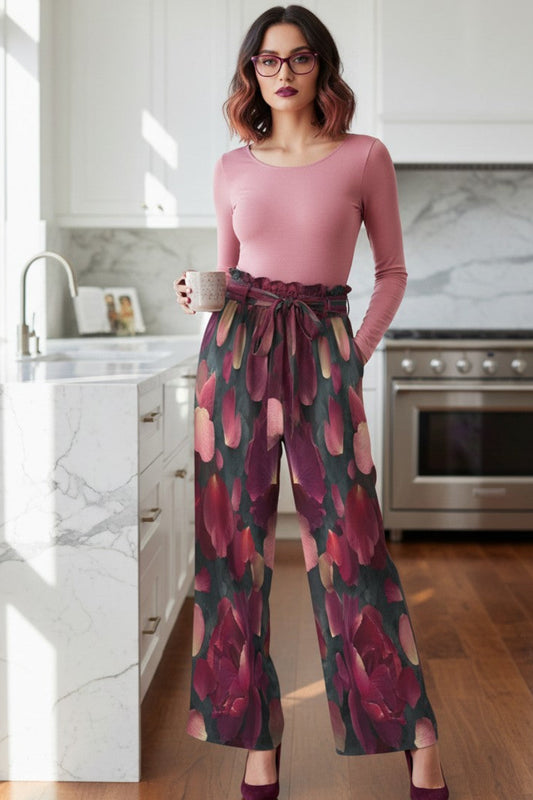 A woman stands in a bright, modern kitchen wearing a dusty pink long-sleeve top and high-waisted, wide-leg trousers with a dark red and green floral pattern. She is holding a mug and wearing glasses.