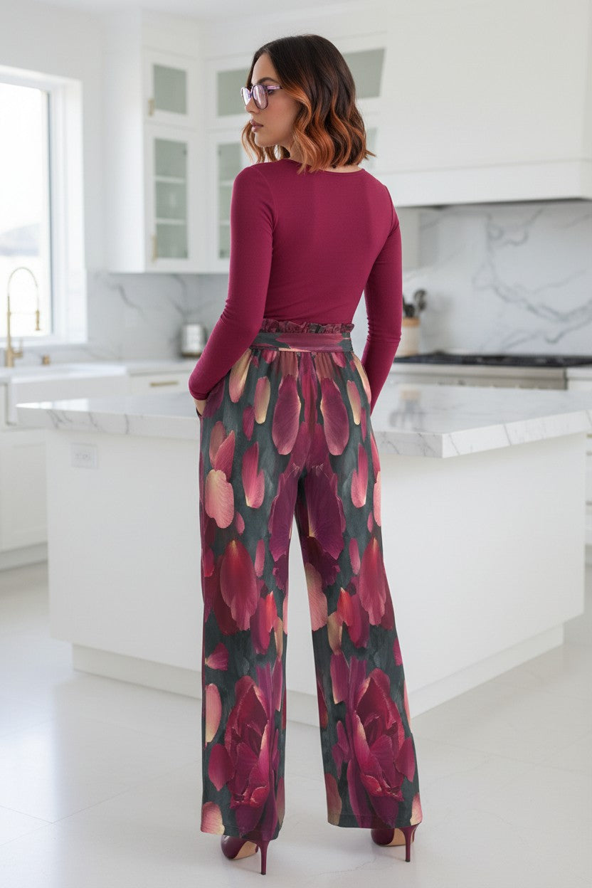 A woman stands in a bright white kitchen, photographed from the back. She is wearing a long-sleeve burgundy top and high-waisted wide-leg trousers with a large floral print in burgundy, pink, and green.