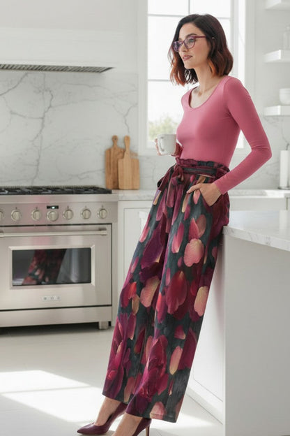A woman wearing a pink long-sleeve top and a dark floral wide-leg skirt leans against a white kitchen island, with a stainless steel oven and marble backsplash in the background.