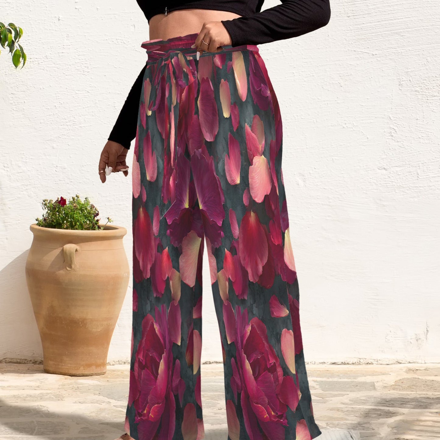 A person wearing high-waisted wide-leg pants with a bold floral print in pink, burgundy, and dark green, styled with a black crop top and clear high-heel sandals against a white wall with potted plants.