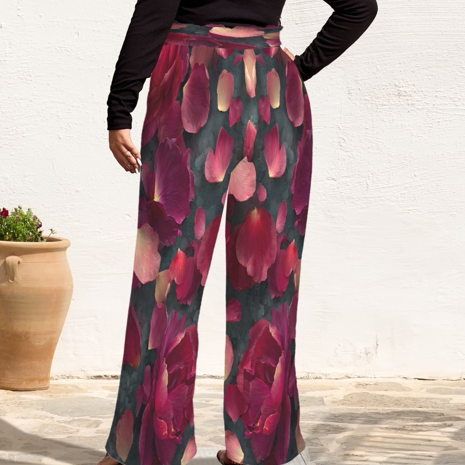 A person wearing high-waisted, wide-leg pants with a dark teal background and large pink and magenta floral print, paired with a black long-sleeve top. The photo is taken outdoors against a white wall with a terracotta planter visible on the left.