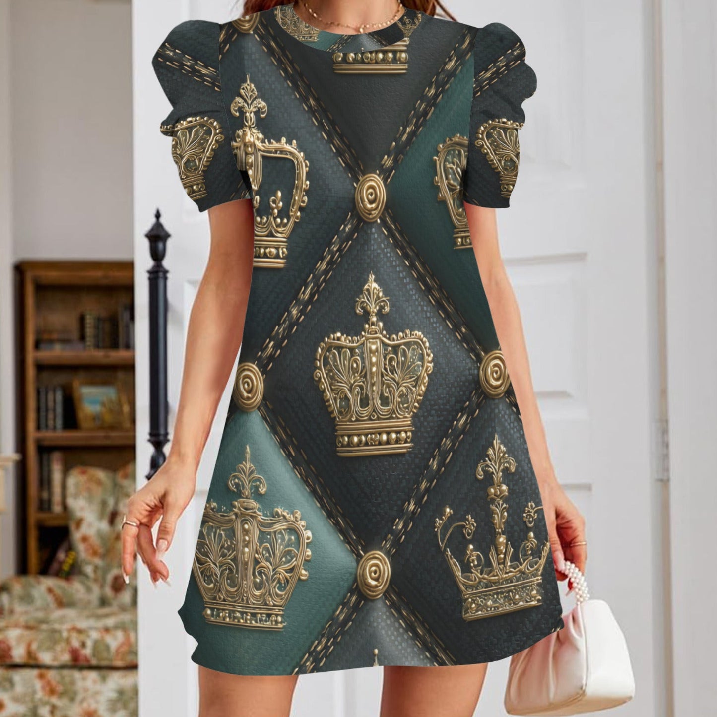 A woman wearing a short-sleeve mini dress with a dark teal and black background and an all-over gold crown pattern, holding a small white handbag in an indoor room setting.