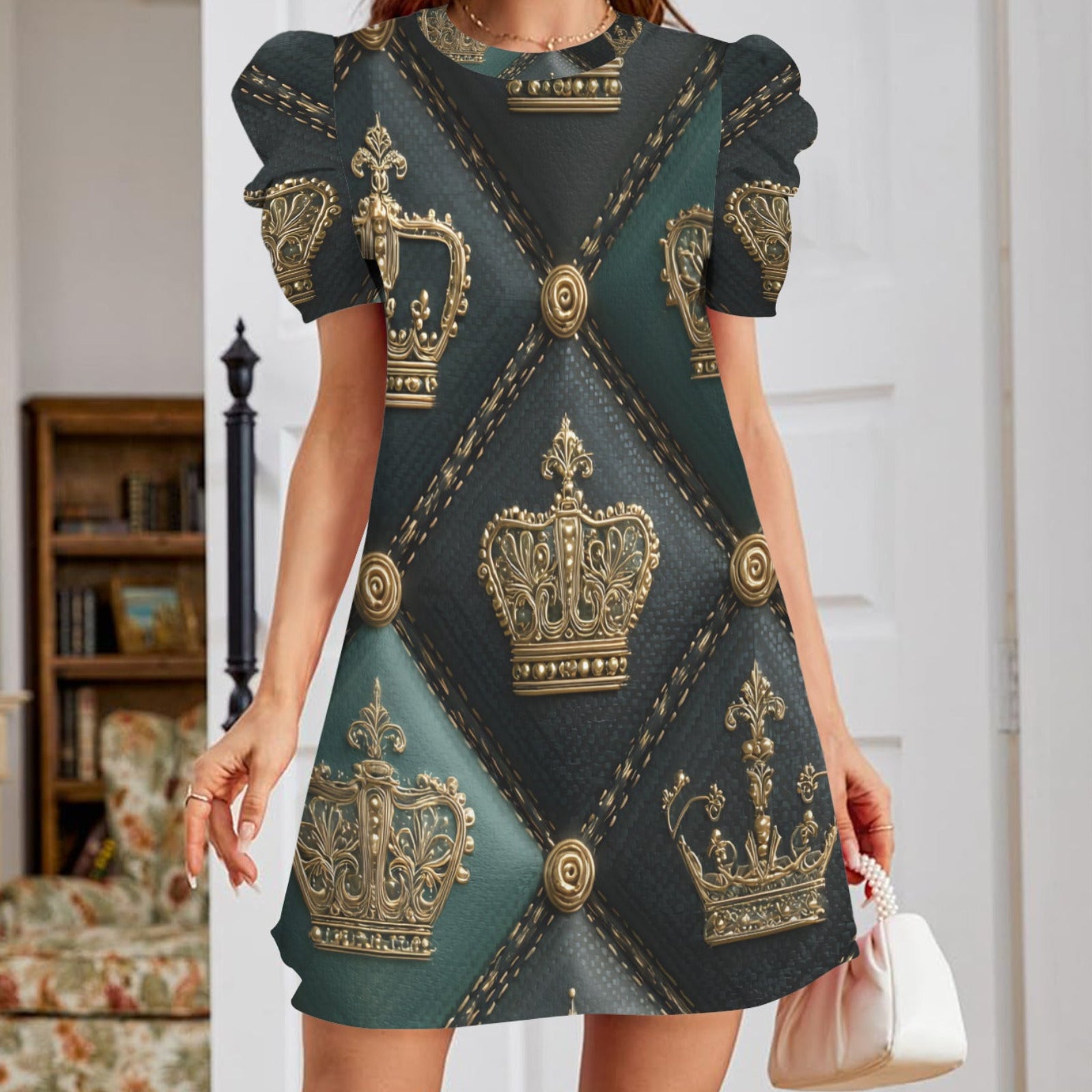 A woman wearing a short-sleeve mini dress with a dark teal and black background and an all-over gold crown pattern, holding a small white handbag in an indoor room setting.