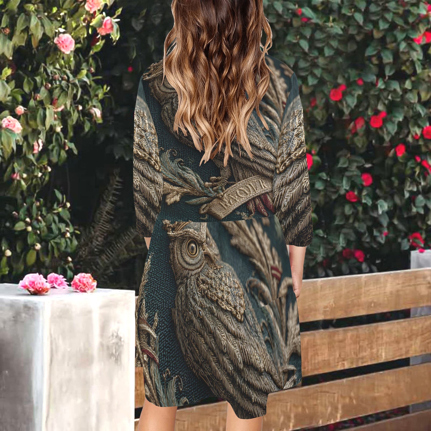 A woman viewed from behind wearing a dark teal and brown owl-patterned kimono cardigan, standing next to a wooden bench in front of a garden with pink flowers.