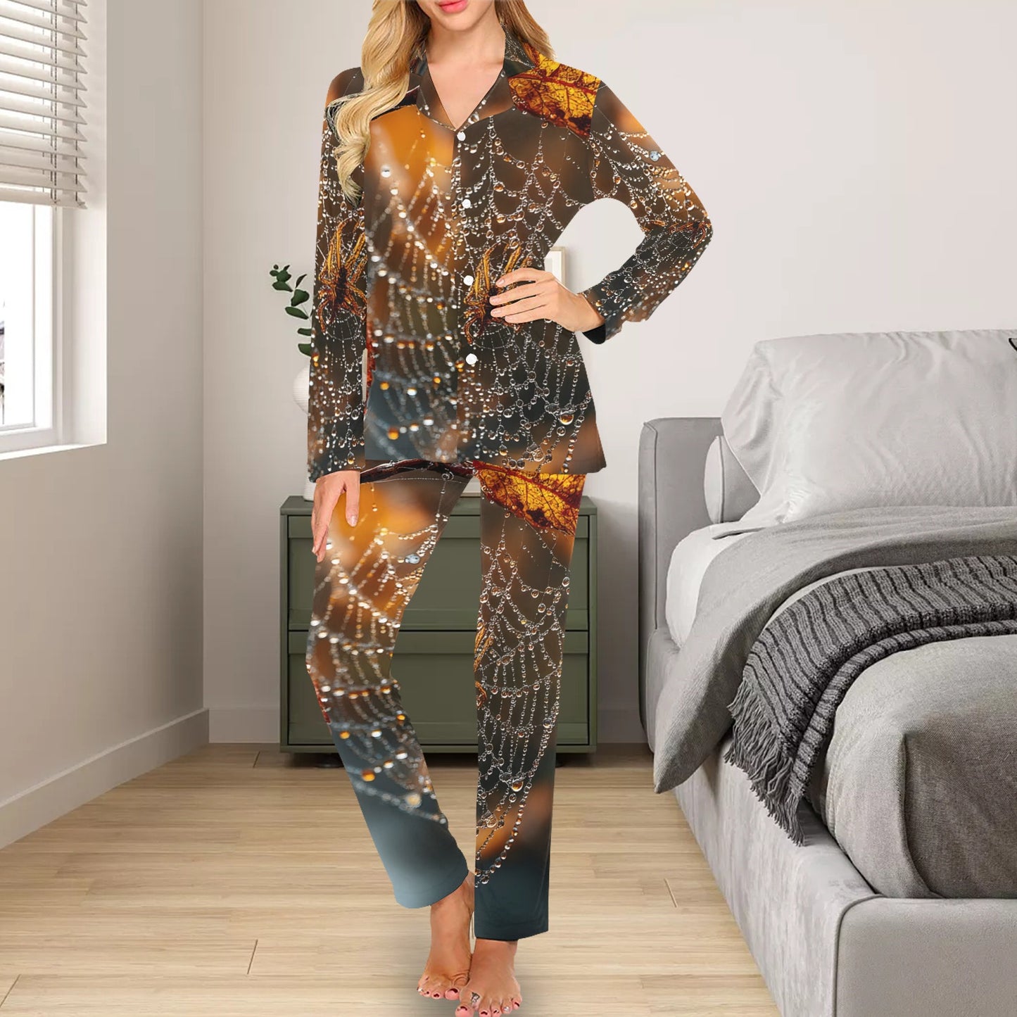 A woman models a two-piece pajama set with a spiderweb and autumn leaf print in brown, orange, and black tones, standing in a bedroom setting.
