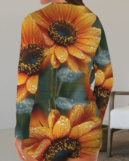 A woman wearing a long-sleeve romper with a vibrant sunflower print, shown from the back in an indoor setting with a white armchair.