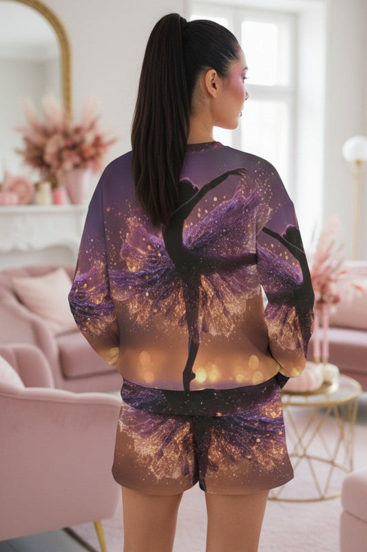 A woman stands with her back to the camera in a bright, pink-toned living room, wearing a matching purple and gold space-themed sweatshirt and shorts set with a black silhouette of a ballerina.
