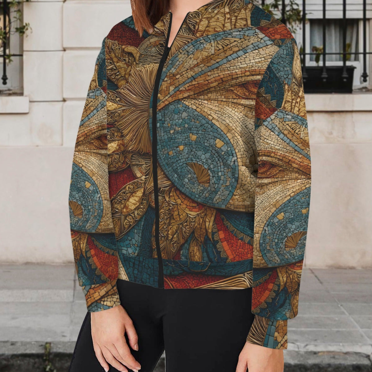 A woman wearing a full-zip bomber jacket with an intricate, vintage-style celestial pattern in teal, gold, rust, and brown, paired with black pants, standing outdoors in front of a light-colored stone building.