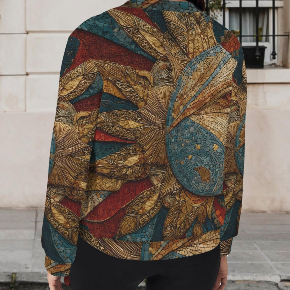 A woman viewed from behind wearing a long-sleeve bomber jacket with an intricate Art Nouveau style pattern in gold, teal, and red, set against a blurred outdoor background of a building facade.