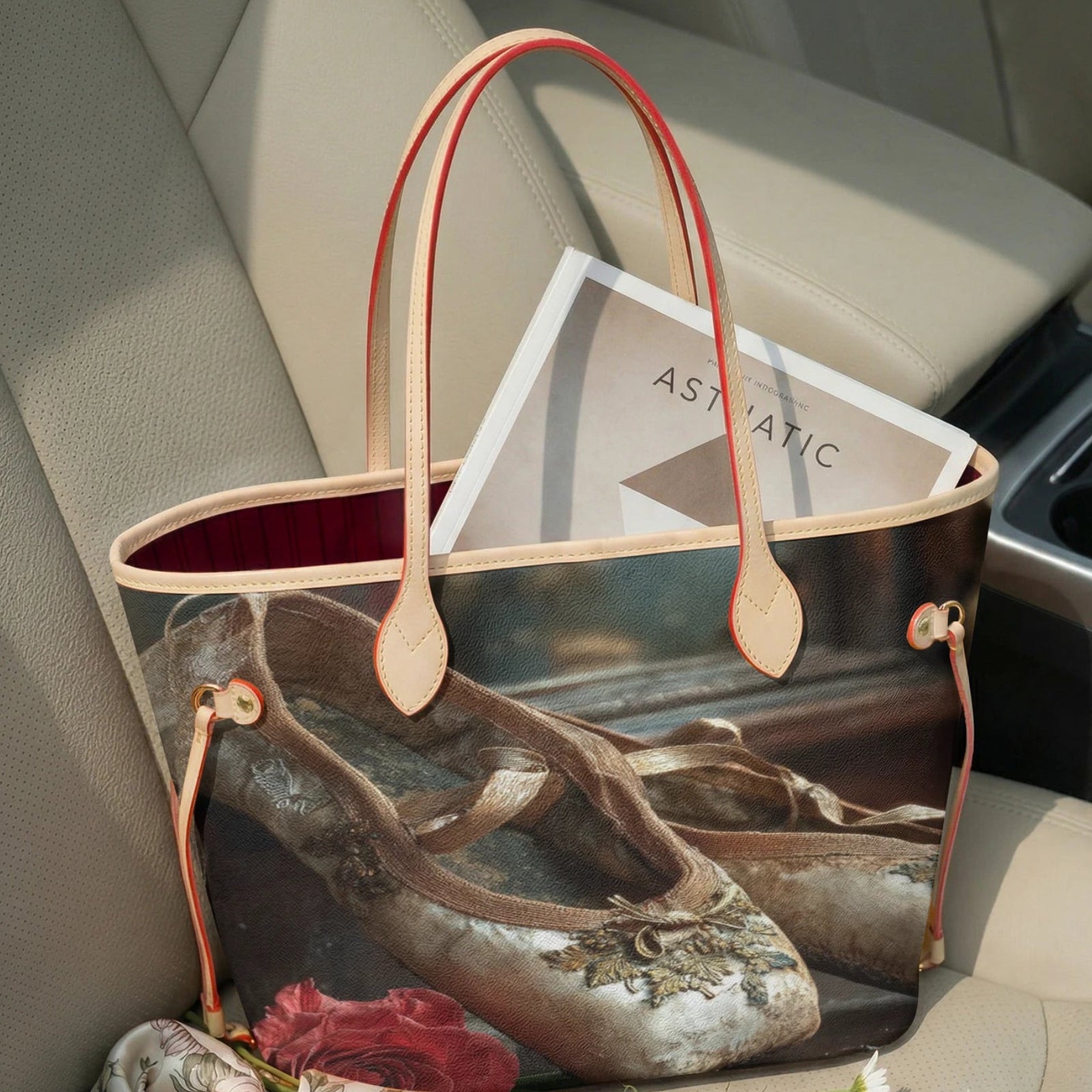 A tote bag with a vintage ballet shoe print and tan leather handles, resting on a beige car seat next to a magazine and flowers.