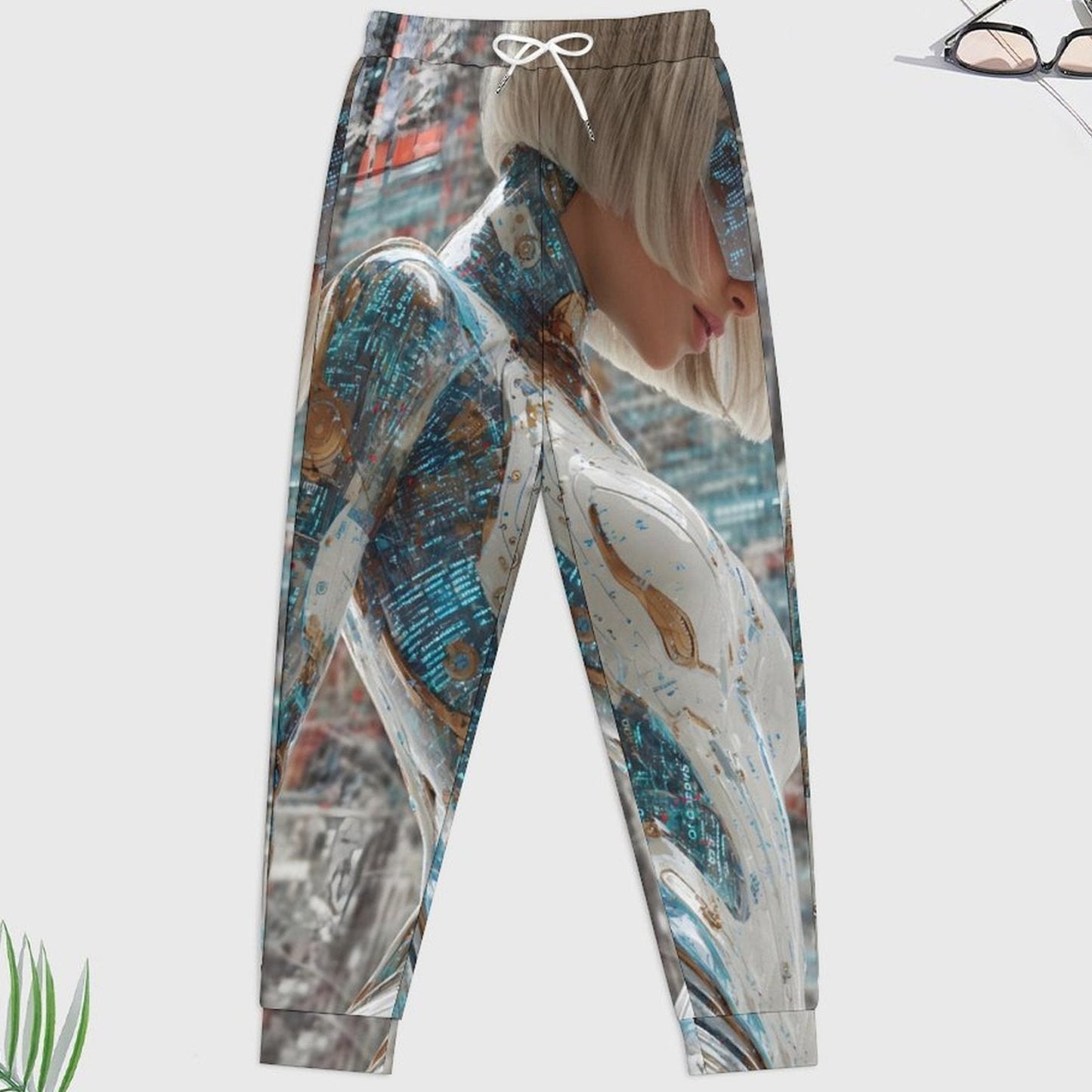 A pair of jogger-style pants with an all-over abstract print featuring a woman in a white dress and blue marbled textures, shown on a light gray background with small decorative plant and eyeglass props.