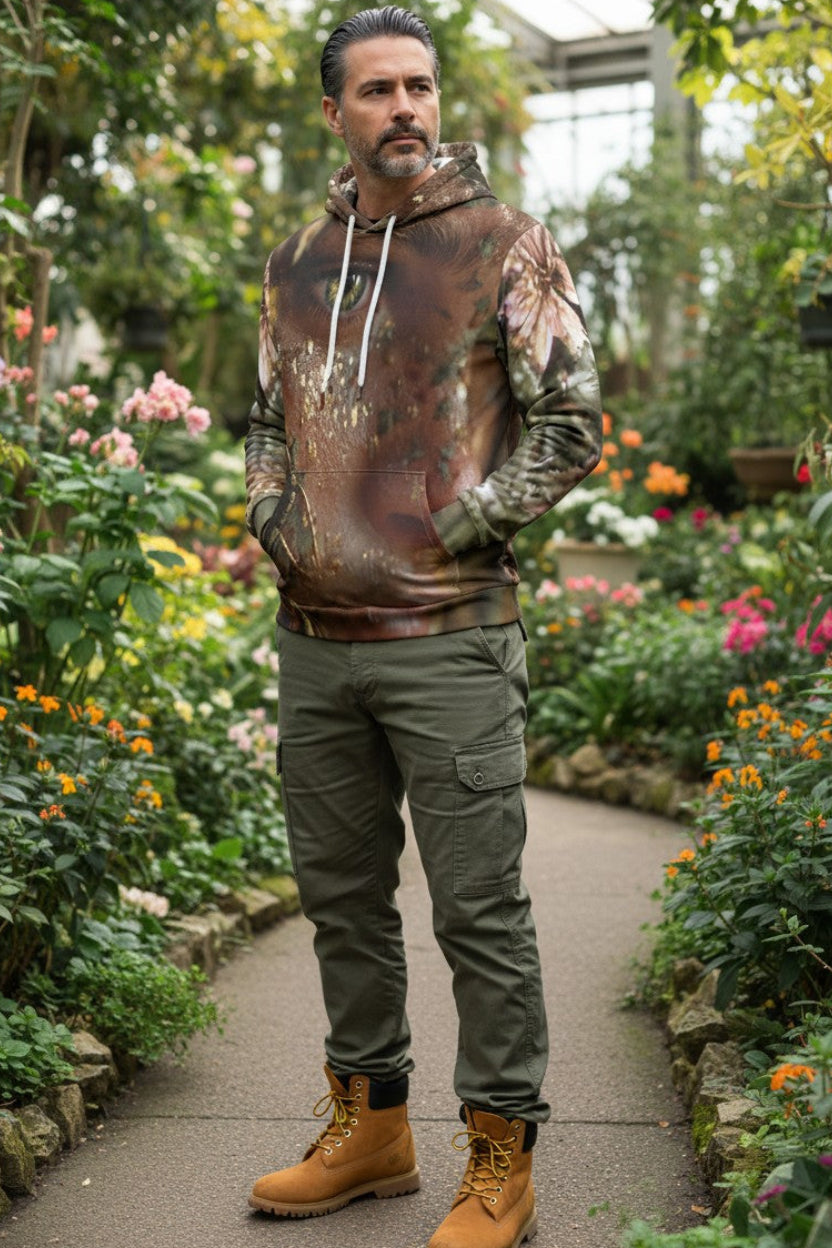 A man stands on a garden path wearing a brown and camouflage patterned hoodie, olive green cargo pants, and tan boots, surrounded by lush green plants and flowers.