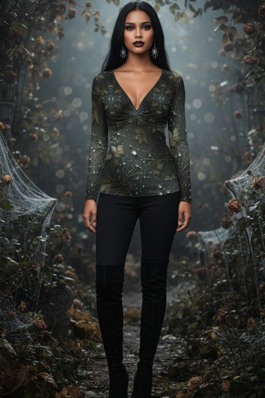 A woman stands in a dark, misty forest path wearing a long-sleeve deep V-neck top with a metallic olive-green and black pattern, paired with black pants and black boots.