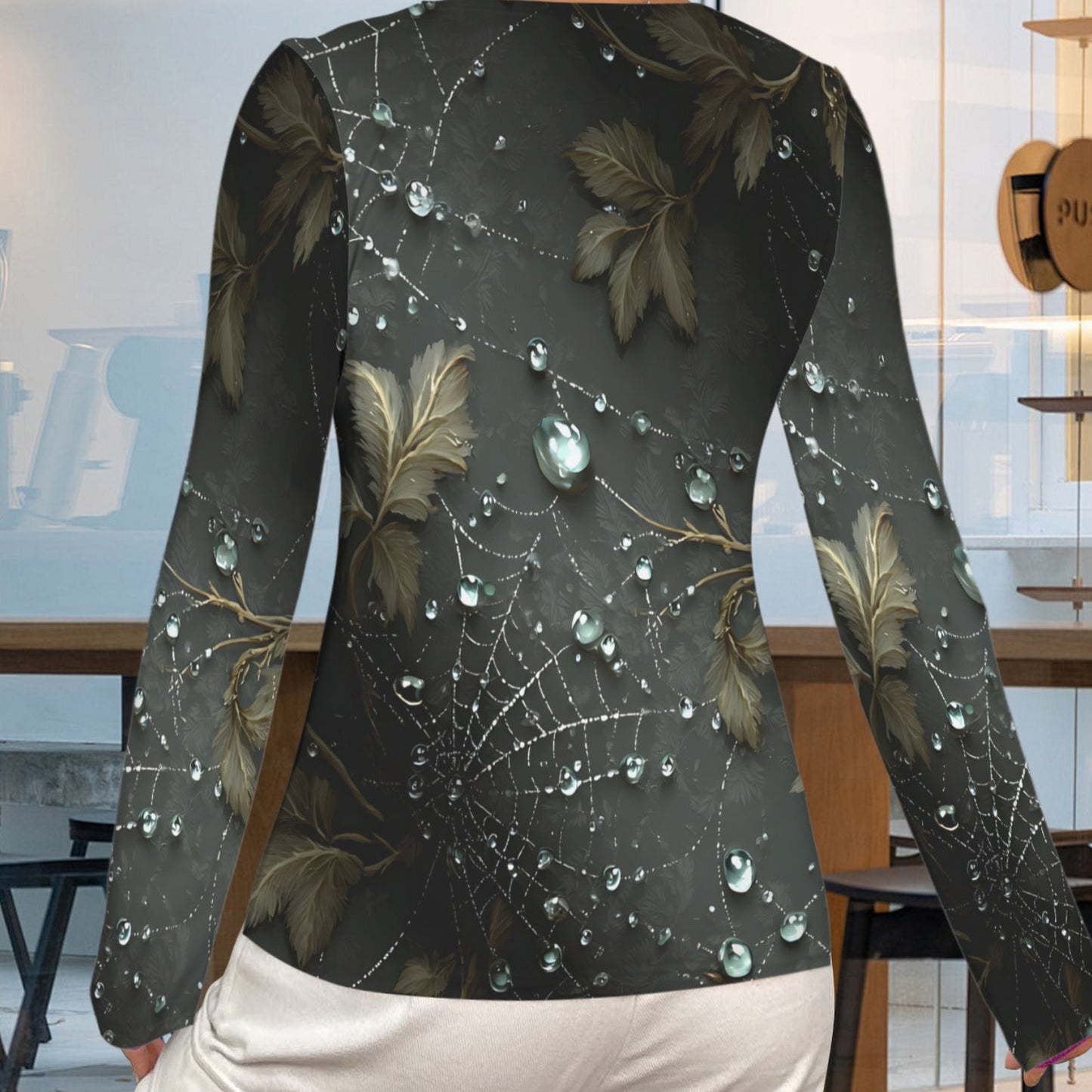 A woman shown from the back wearing a long-sleeve top with a dark gray and black spiderweb and leaf pattern, featuring realistic water droplets, paired with white pants in a modern cafe setting.