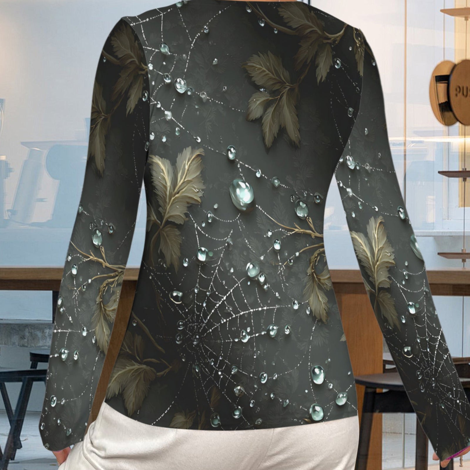 A woman shown from the back wearing a long-sleeve top with a dark gray and black spiderweb and leaf pattern, featuring realistic water droplets, paired with white pants in a modern cafe setting.