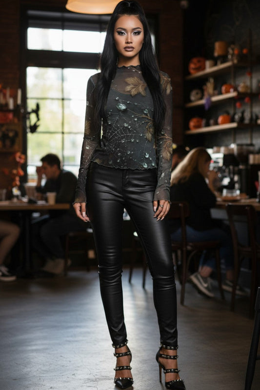 A woman modeling a dark sheer floral long-sleeve top with sequined detailing, paired with black faux-leather pants and black studded high heels, standing in a dimly lit cafe interior.
