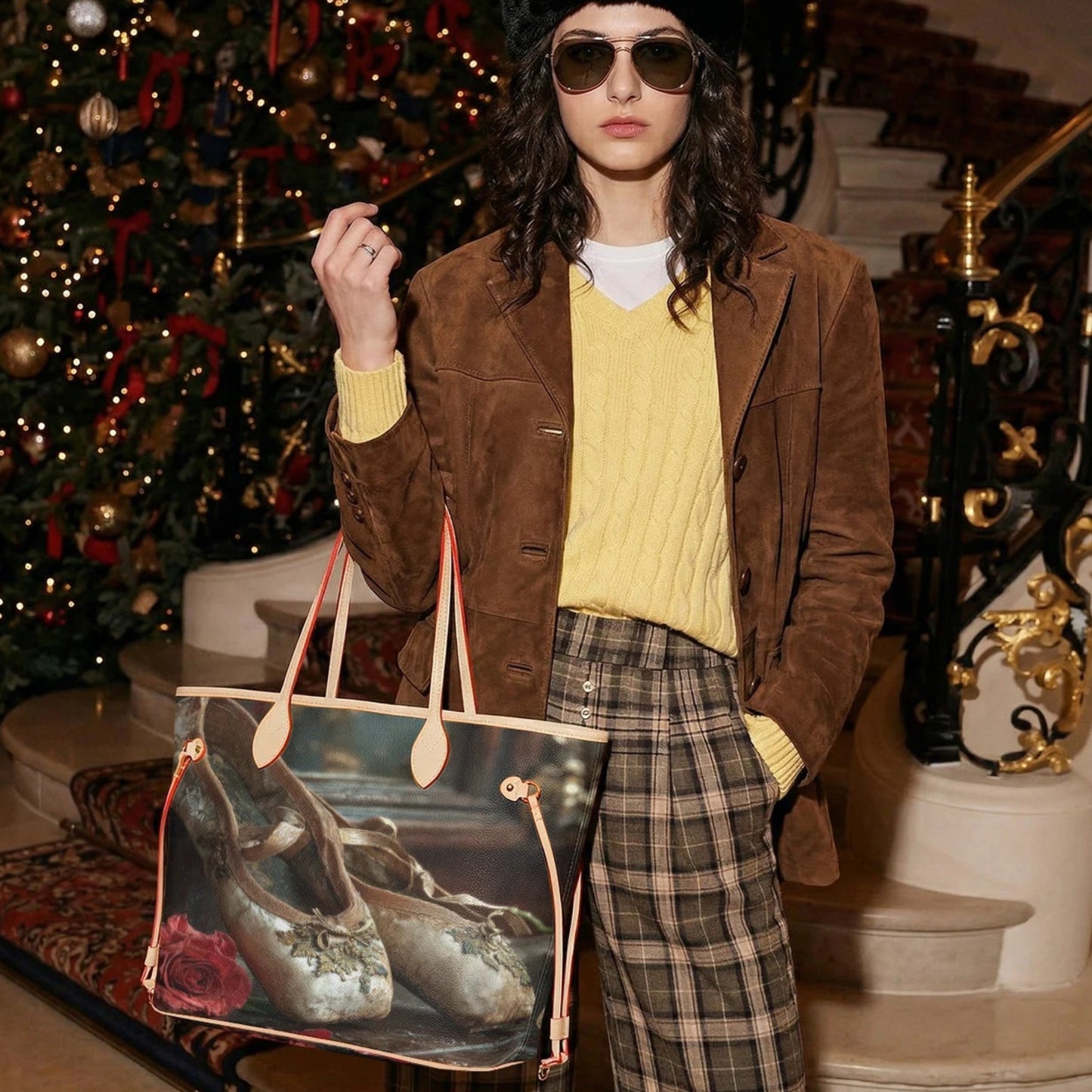 A model stands in an elegant indoor setting with a staircase and a decorated Christmas tree, holding a large canvas tote bag. The bag features a photographic print of a pair of silver high-heeled shoes and a red rose, framed by beige canvas borders and beige leather handles.